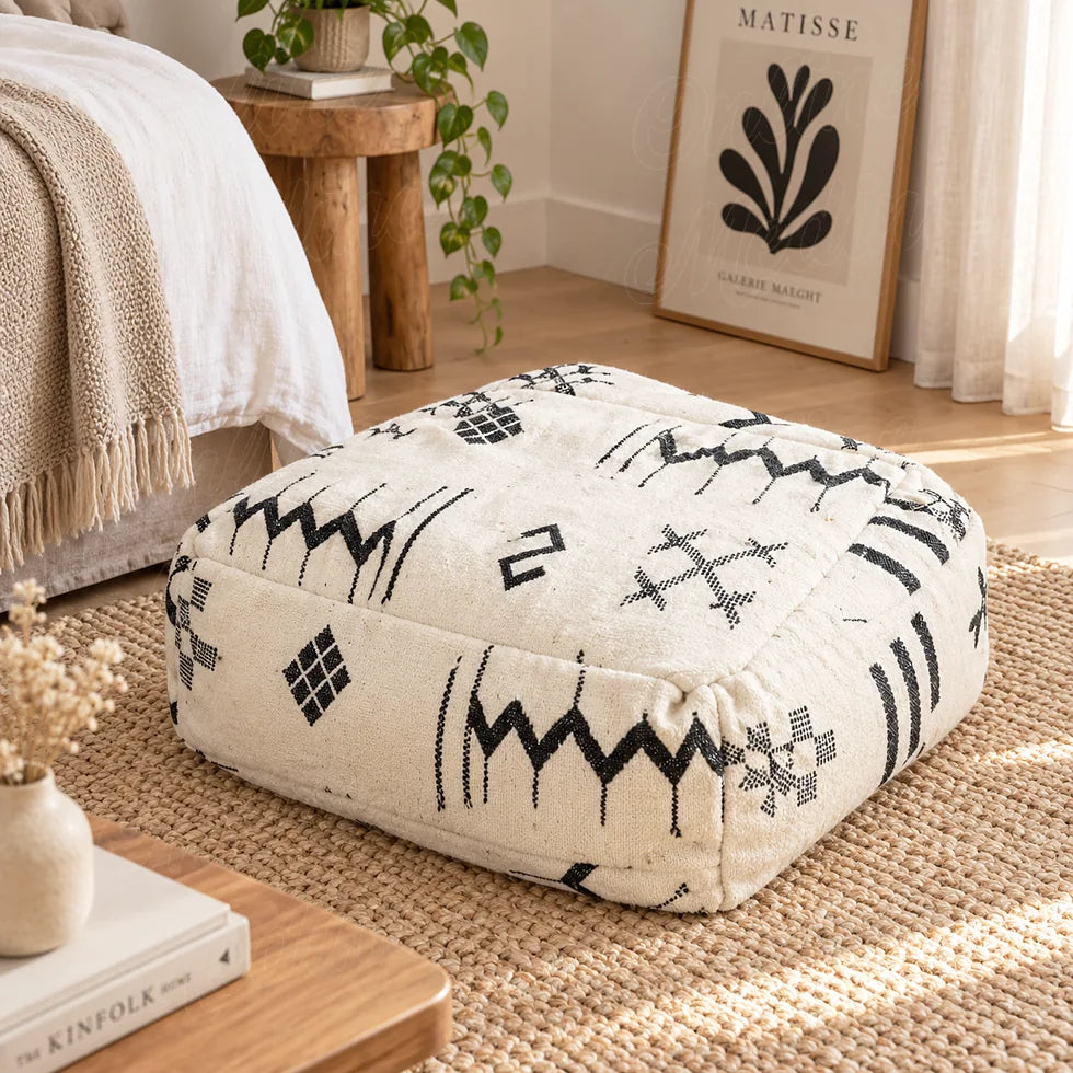 Puf de lana marfil con zigzags y rombos negros junto a una cama, sobre una alfombra beige en un dormitorio luminoso