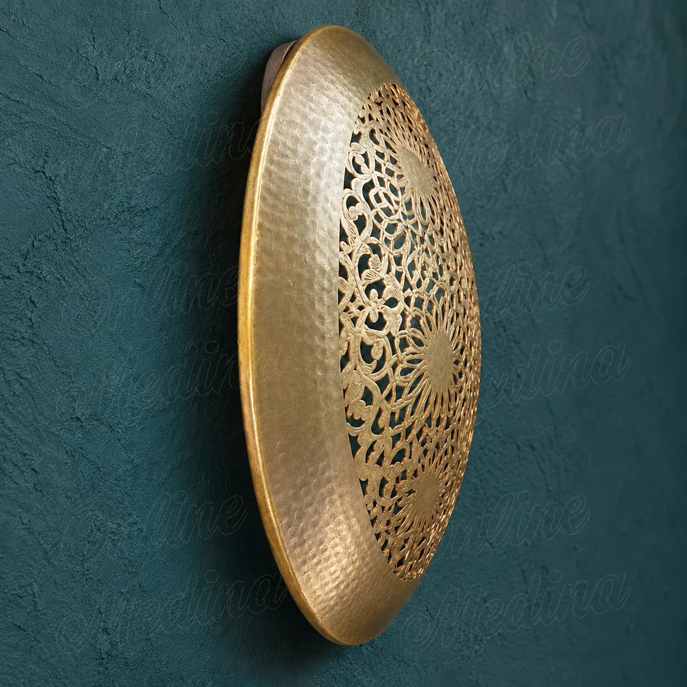 Side view of a round golden brass wall sconce, showing its domed profile and hammered rim against a blue backdrop.