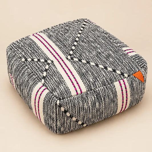 Pouf carré en laine chinée noire et crème, avec bandes fuchsia et détail orange, sur fond beige