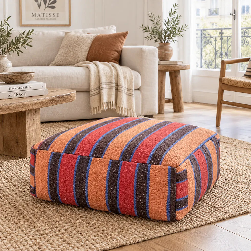 Wool pouf with red, orange, brown and blue stripes on a beige rug in front of a light-colored sofa