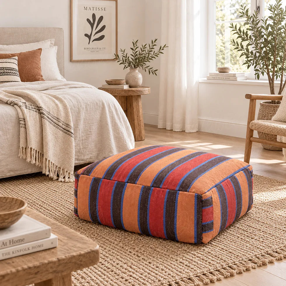 Wool pouf with red, orange, brown and blue stripes at the foot of a bed in a bright bedroom