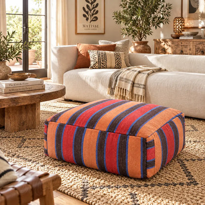 Red, orange, brown and blue striped wool pouf in front of a cream sofa beside a round wooden table