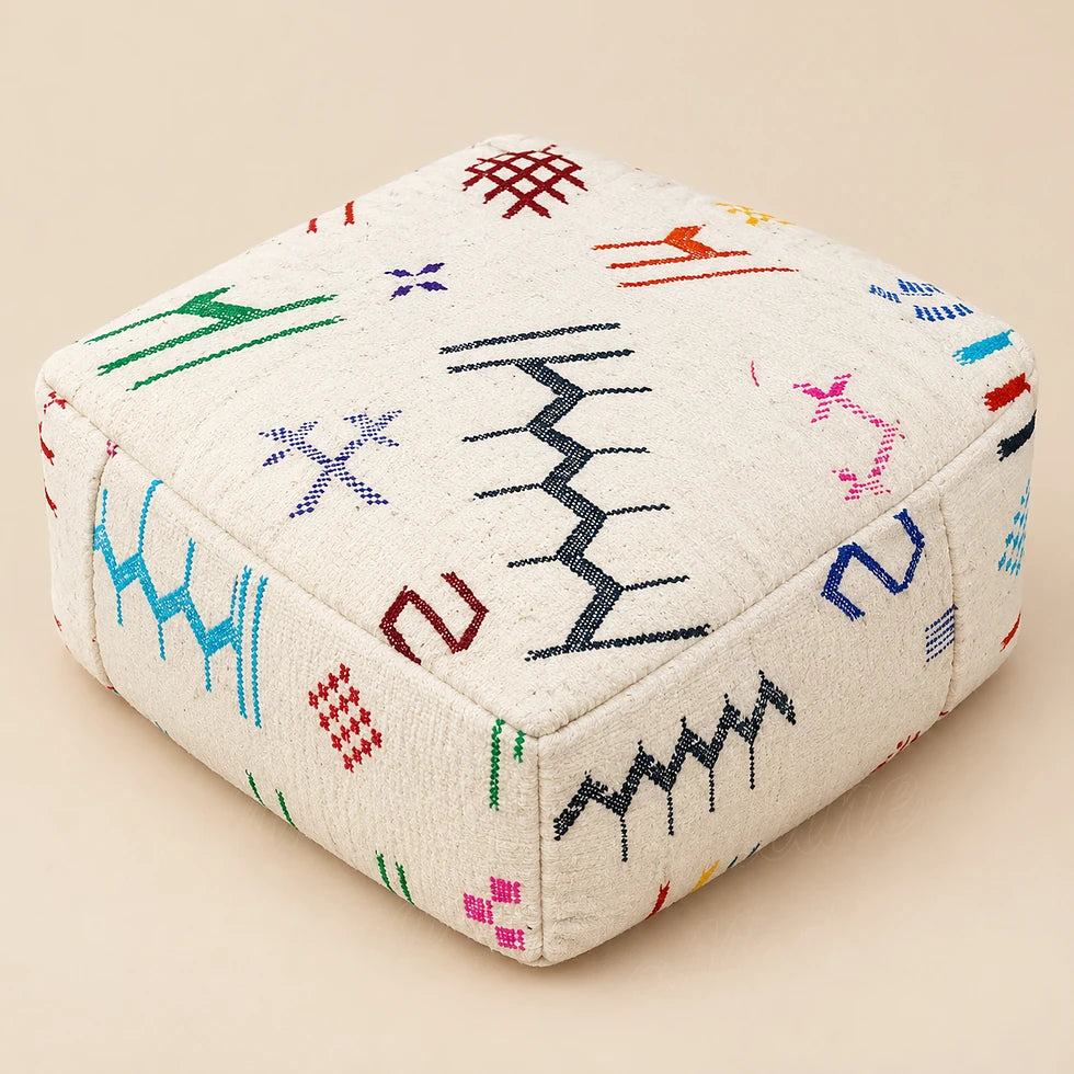 Square ivory wool pouf with a black zigzag and colorful motifs, shown at an angle on a beige background