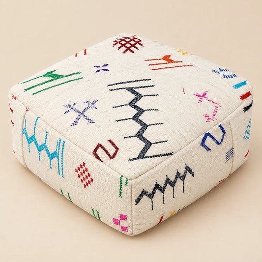 Square ivory wool pouf with a black zigzag and colorful motifs, shown at an angle on a beige background