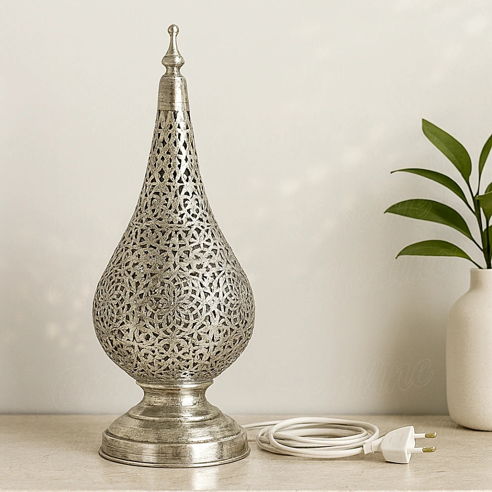 A silver Moroccan tear-shaped table lamp with intricate floral patterns, set on a beige table next to a green plant in a contemporary interior.