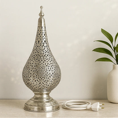 A silver Moroccan tear-shaped table lamp with intricate floral patterns, set on a beige table next to a green plant in a contemporary interior.