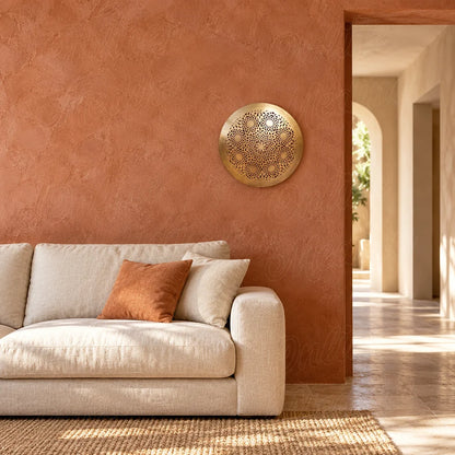 Round golden brass wall sconce with openwork floral rosettes, above a sofa on a terracotta wall.