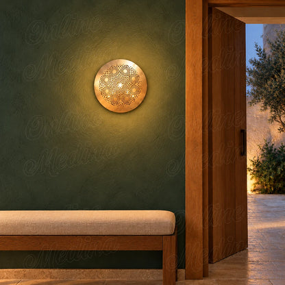 Round golden brass wall sconce with openwork floral rosettes, illuminated with a warm halo on a green wall.