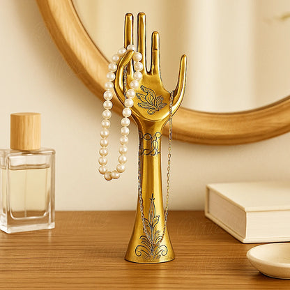 Brass hand-shaped jewelry holder with engraved motifs holding a pearl necklace and gold chain on a wooden vanity.