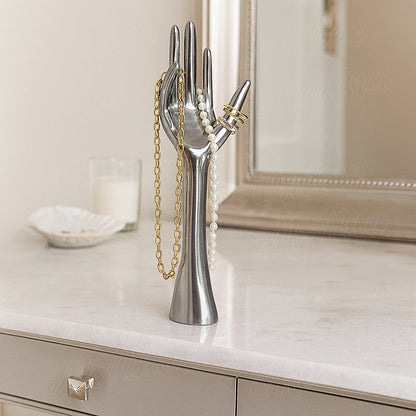 Aluminum hand jewelry holder displaying a gold chain, pearl necklace, and rings on a marble vanity with mirror.