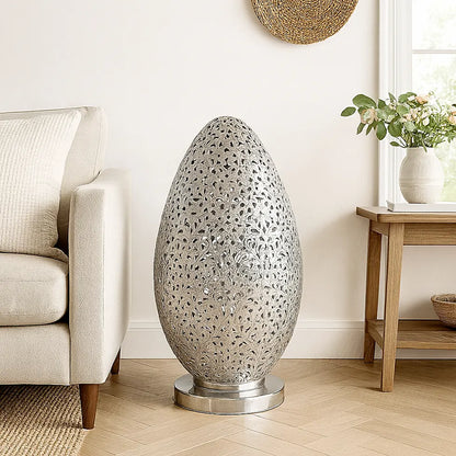 Large Moroccan silver egg-shaped lamp placed on the floor beside a beige sofa in a bright modern living room with wooden furniture.