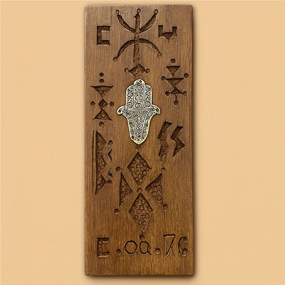 Front view of a handcrafted Moroccan cedar wood wall amulet with Amazigh carvings and a silver Fatima hand on a beige background.
