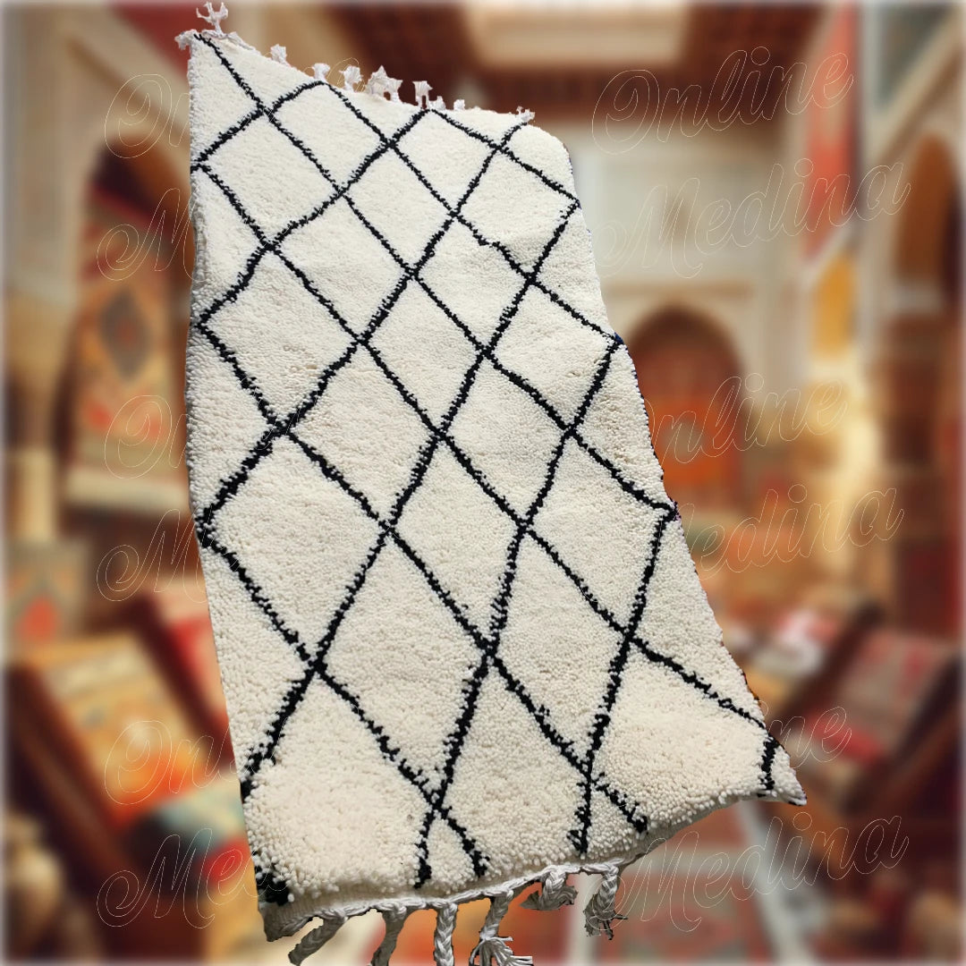 Tapis Beni Ouarain marocain fait main en laine blanche avec motifs noirs en losanges, présenté dans un souk artisanal pour une décoration authentique.