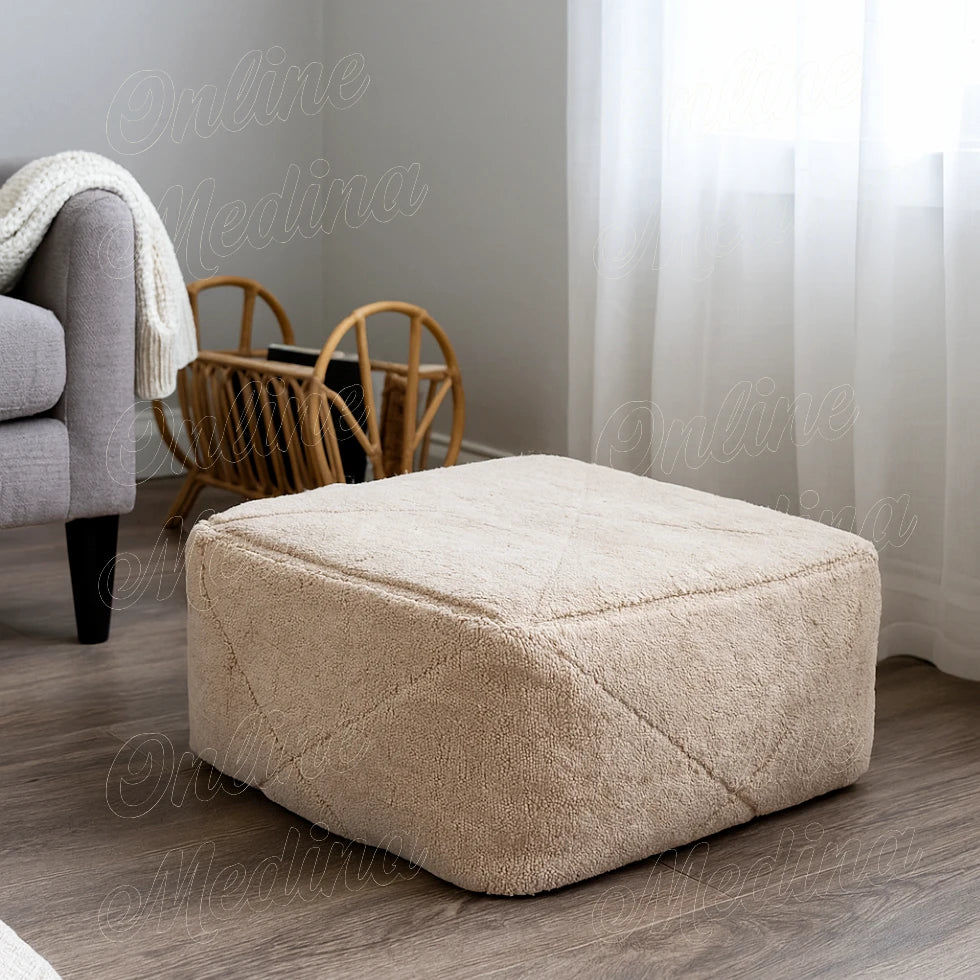 Handwoven Moroccan pouf made of beige wool, placed in a bright minimalist living room with wooden furniture and white curtains.
