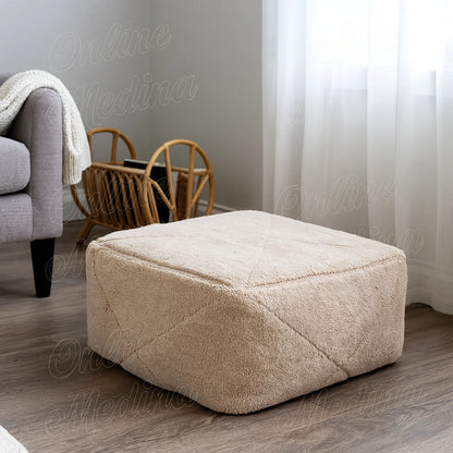 Handwoven Moroccan pouf made of beige wool, placed in a bright minimalist living room with wooden furniture and white curtains.